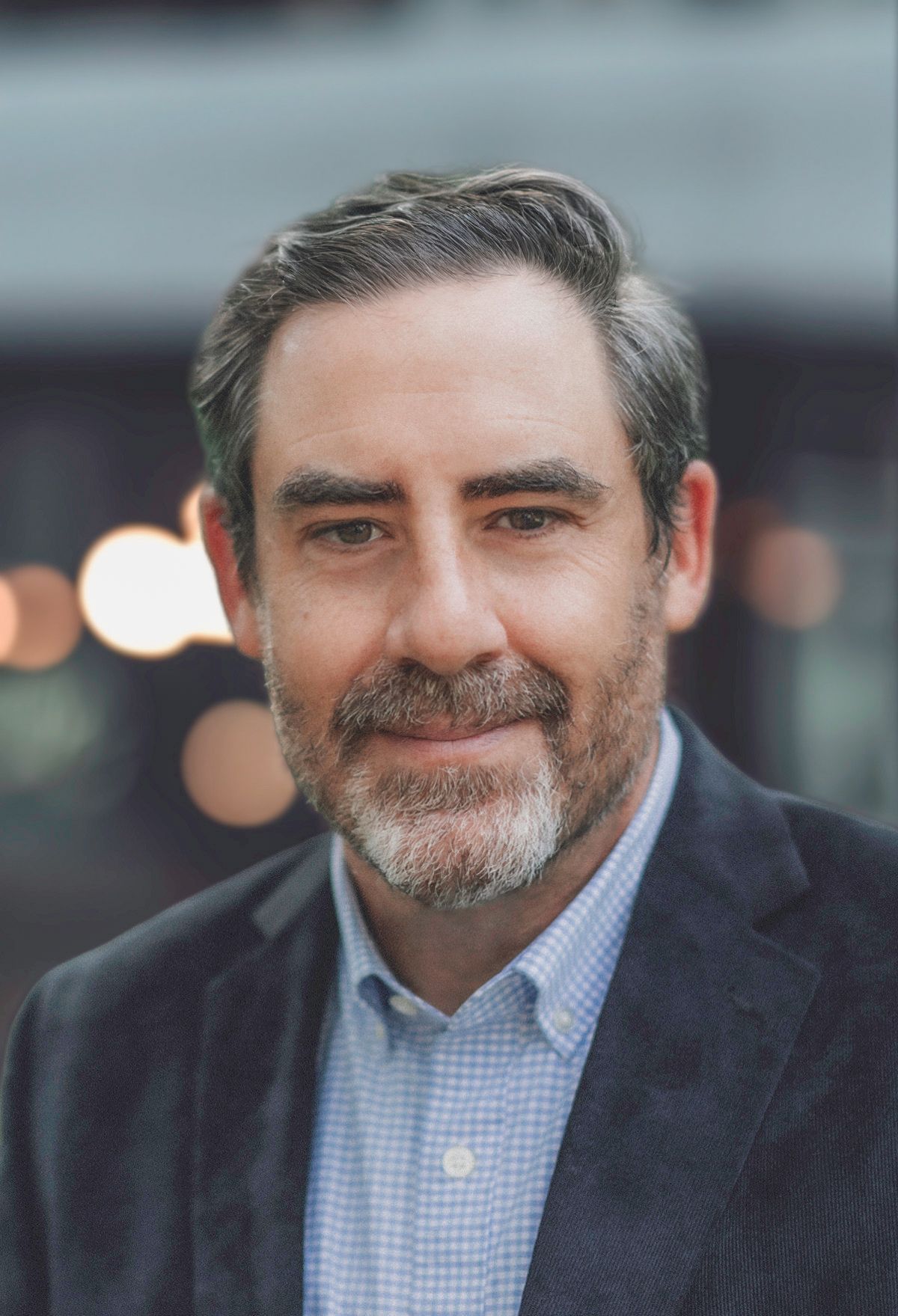 Professional headshot portrait of someone in a navy blazer and light blue checkered shirt against a blurred background.