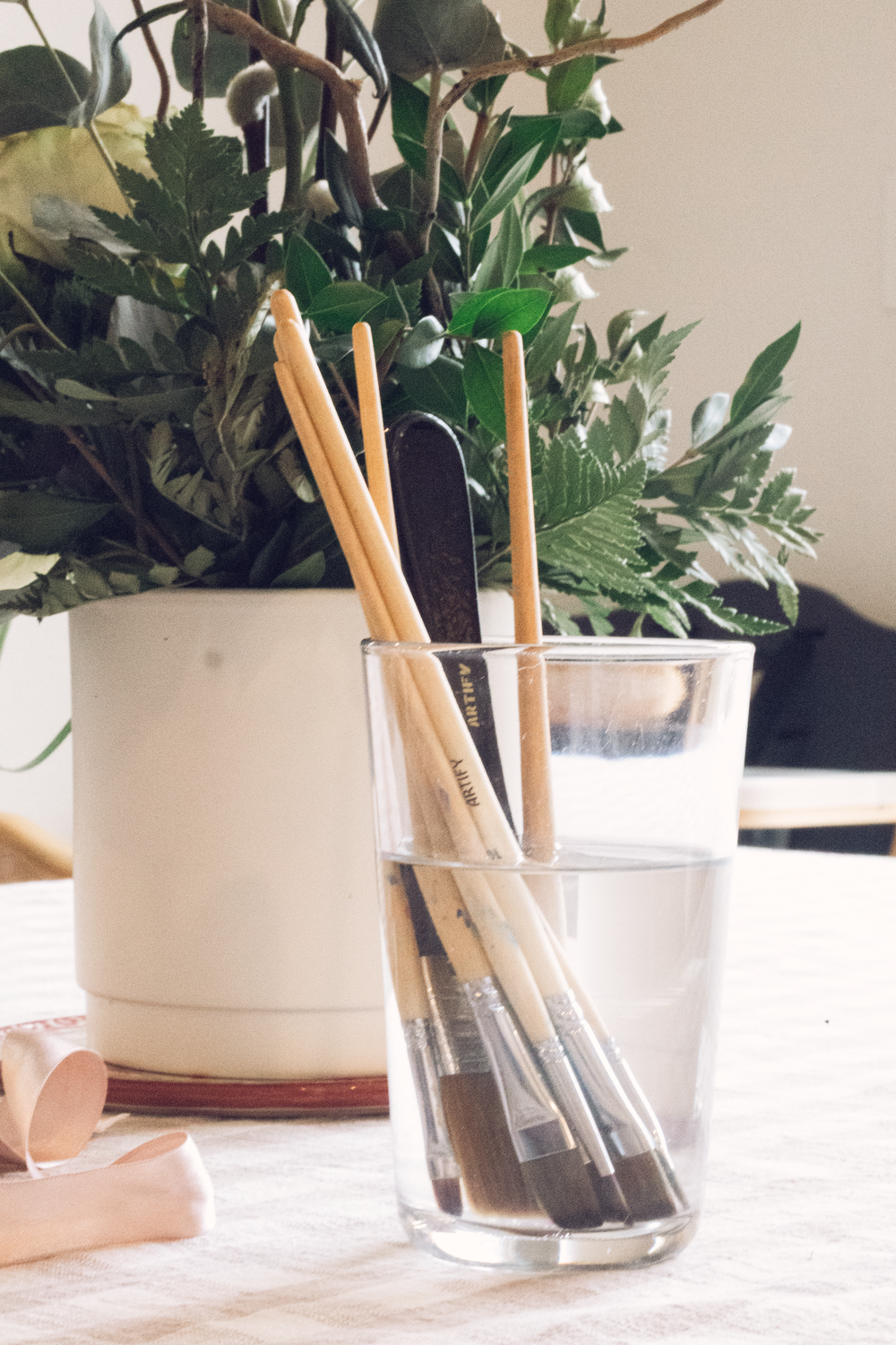 Paint brushes soaking in a clear glass next to a white vase with green foliage on a table.