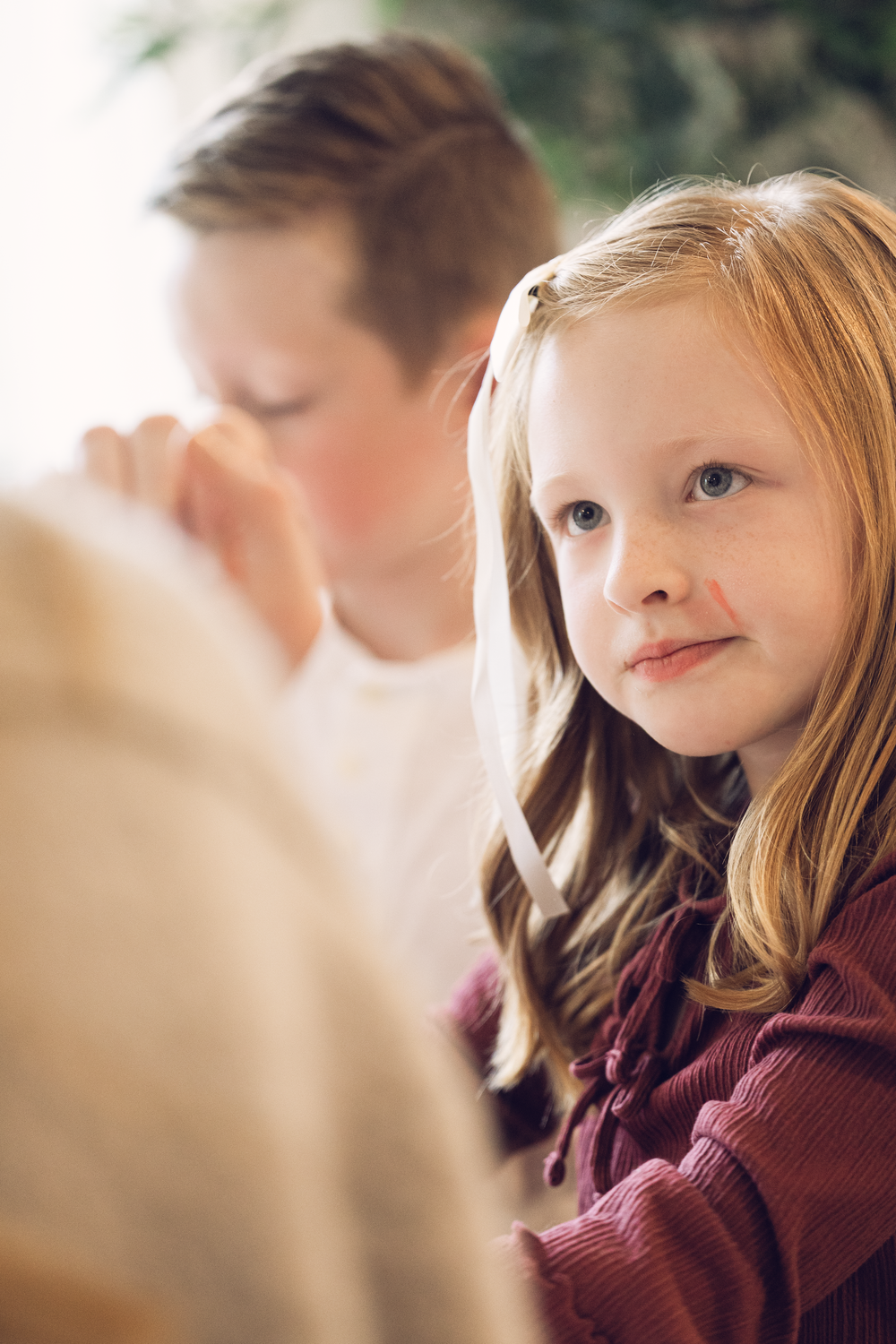Young girl with long blonde hair looking at camera while boy stands behind her in soft light.