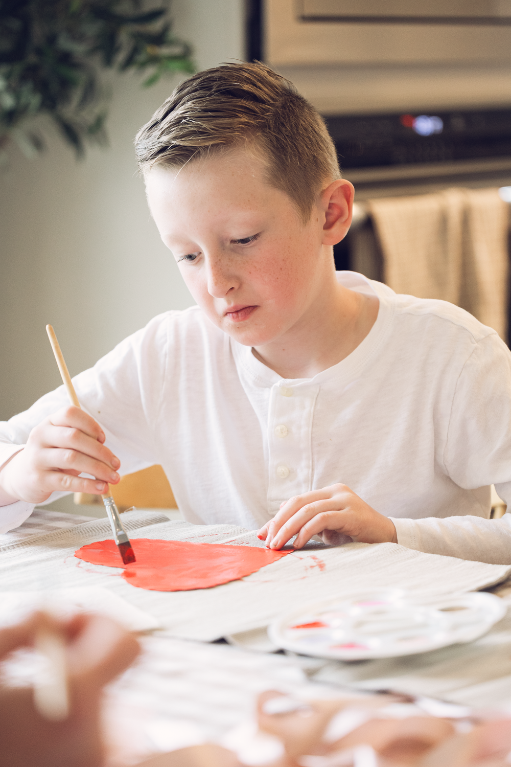 Young boy in white shirt carefully painting a red heart shape with a brush at a table.
