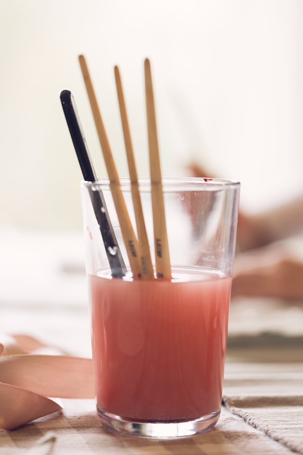 Glass jar with red-tinted water holding wooden chopsticks and paint brushes on a table.