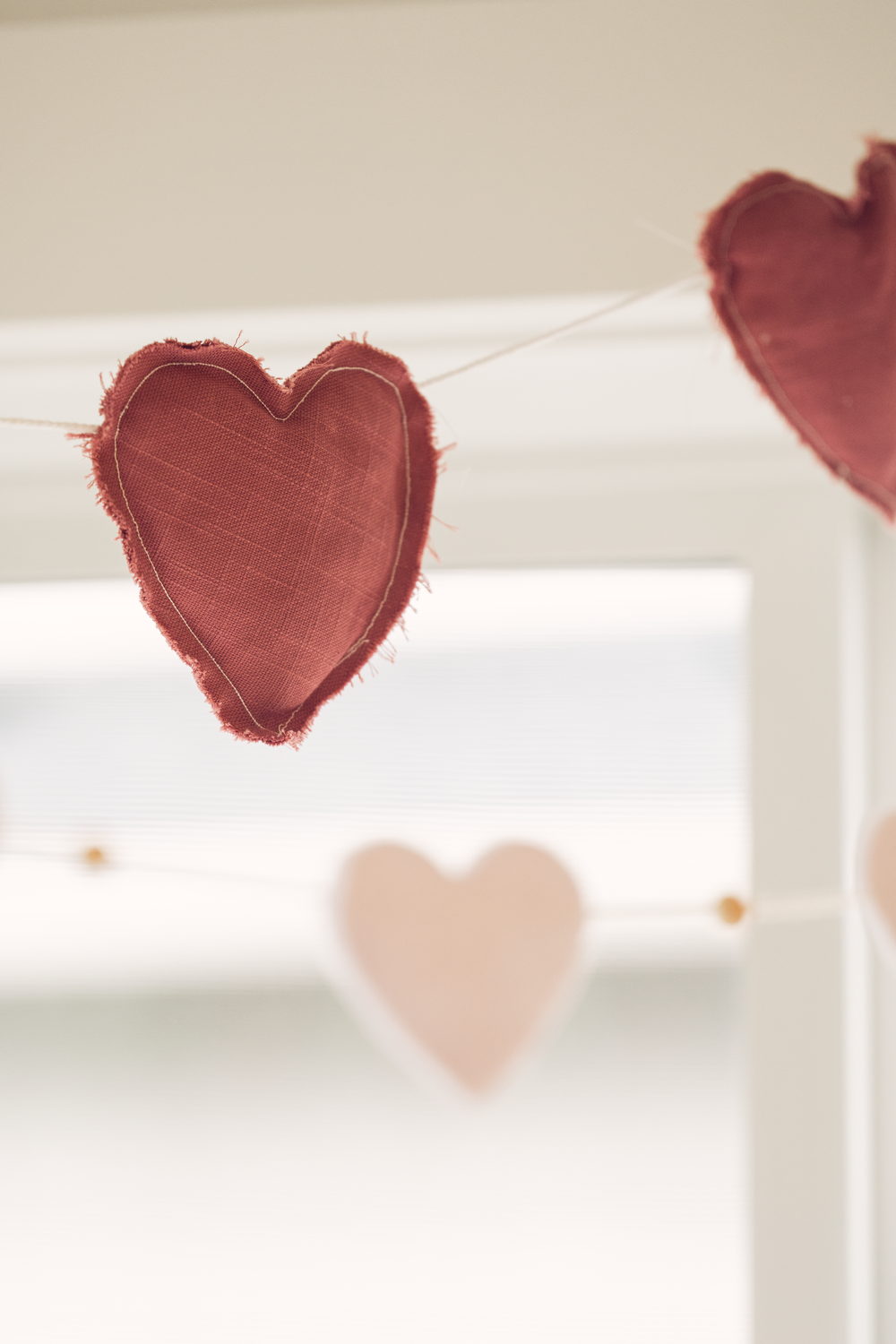 Handmade fabric heart garland in red and pink hanging near a bright window as holiday decor.