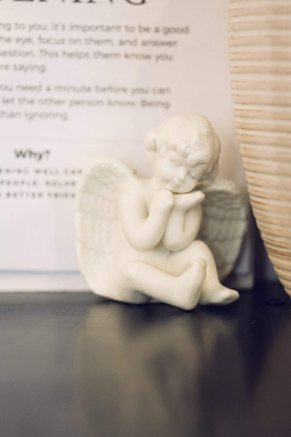 White ceramic cherub angel figurine sitting pensively beside a wicker basket on a dark shelf.