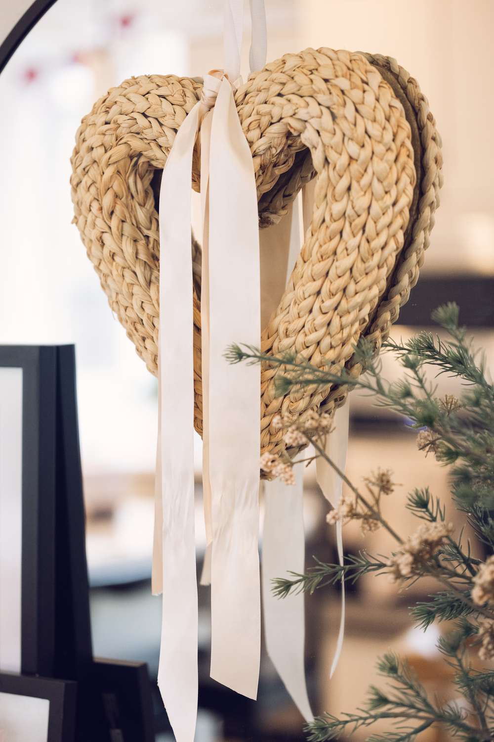 Heart-shaped woven rope wreath with white ribbon hanging near pine branches and a dark frame.