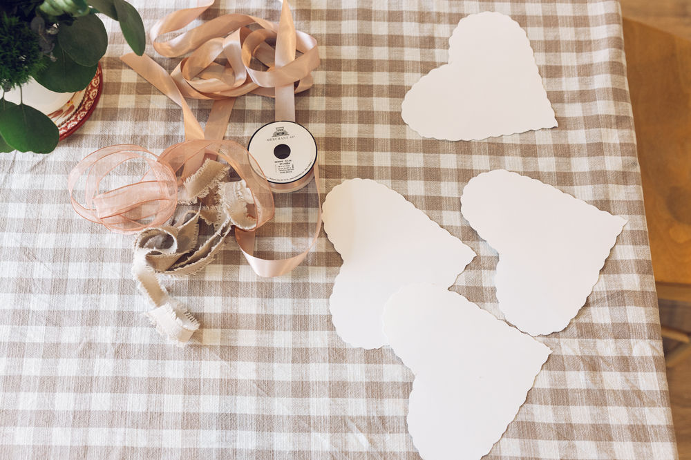 White paper heart cutouts with pink ribbon spool on a beige gingham tablecloth for crafting.