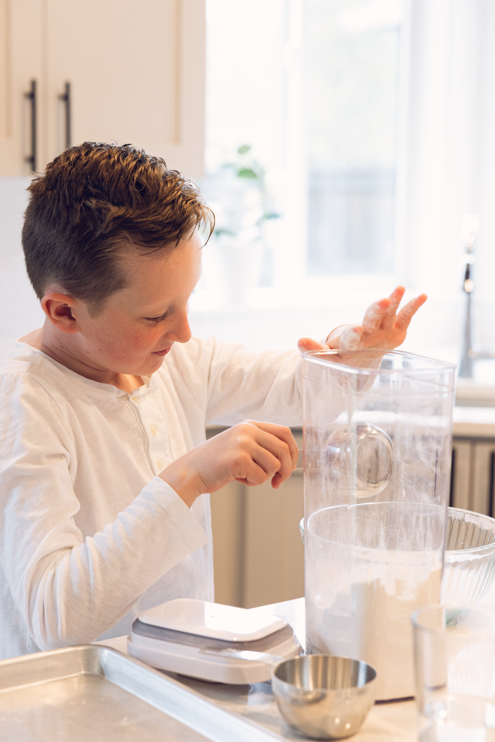 Young boy in white shirt using a blender jar on a kitchen scale in a bright white kitchen.