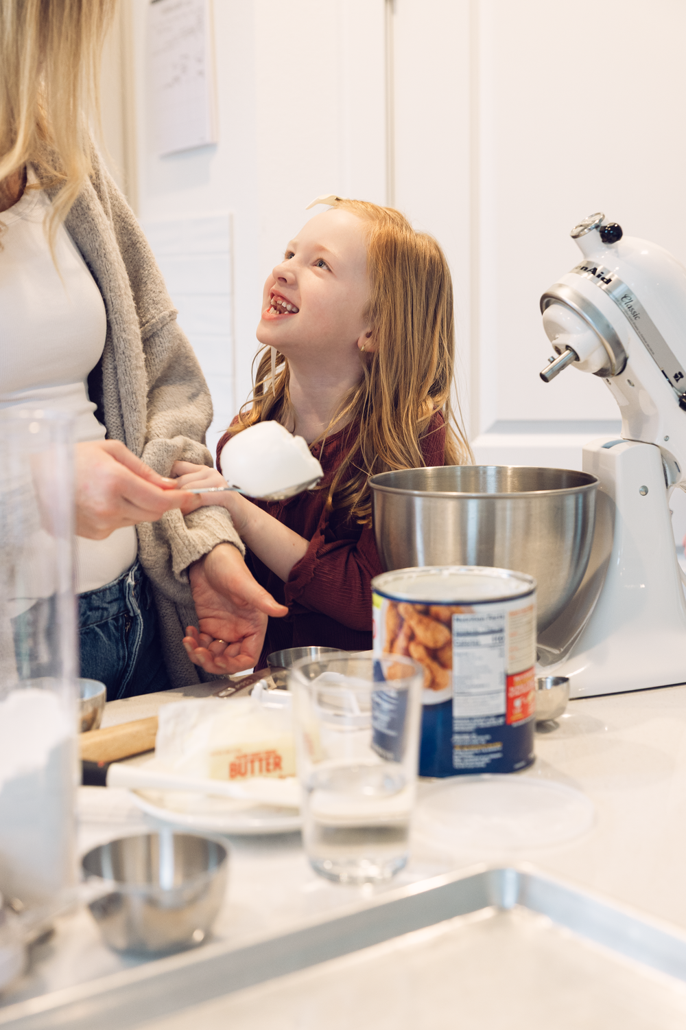 Happy red-haired girl baking with mother near a stand mixer in a bright white kitchen.