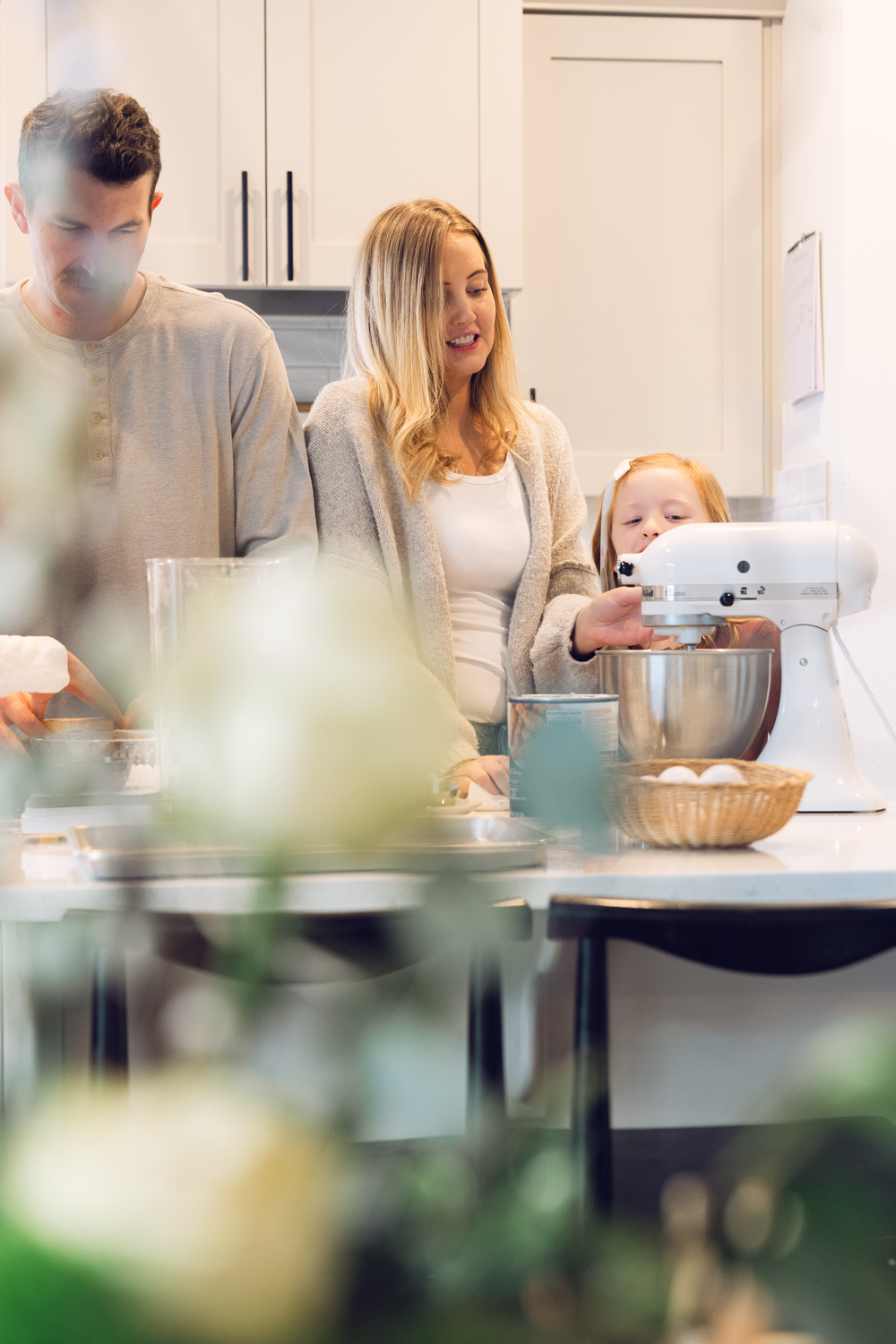 Family of three baking together in a modern white kitchen with a stand mixer and ingredients.