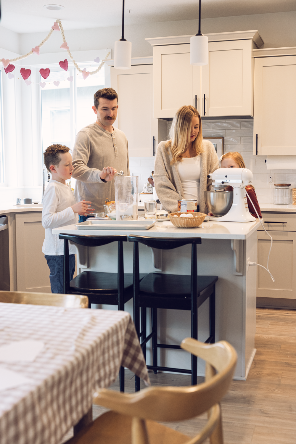 Family of four baking together in a modern white kitchen with heart garland decorations.