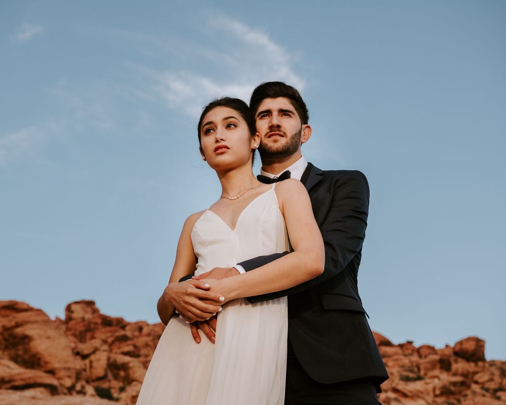 Couple in wedding attire embracing against red rock canyon backdrop under blue sky.