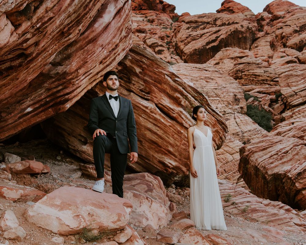Bride and groom posing separately against massive red sandstone rock formations in desert.