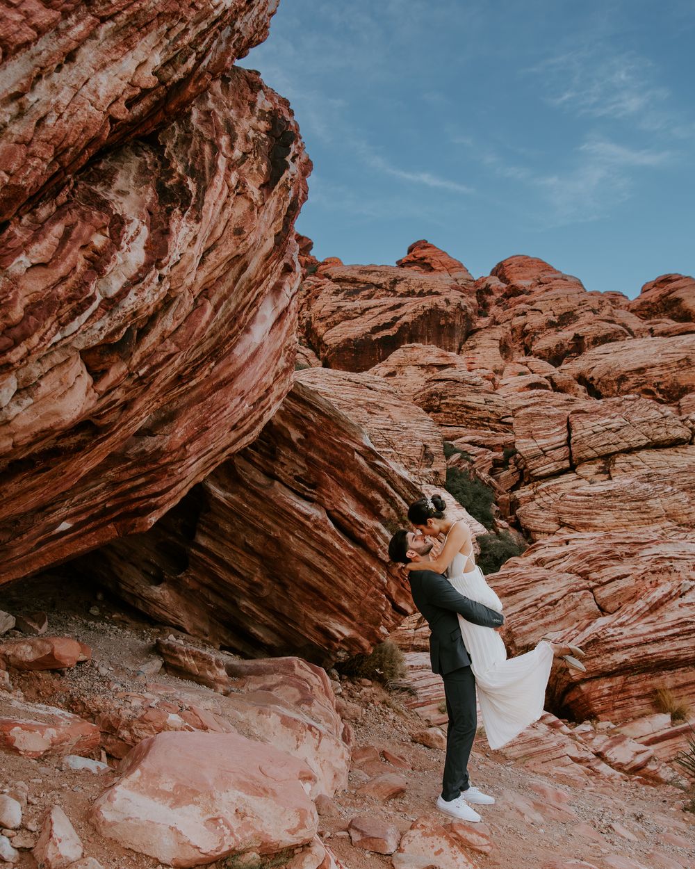 Groom lifting bride in white gown against layered red rock canyon formations outdoors.