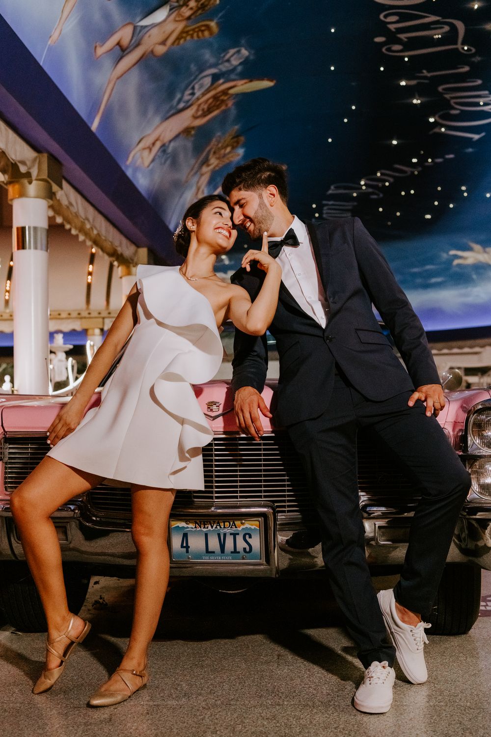 Couple in wedding attire leaning on pink Cadillac with Elvis license plate indoors.