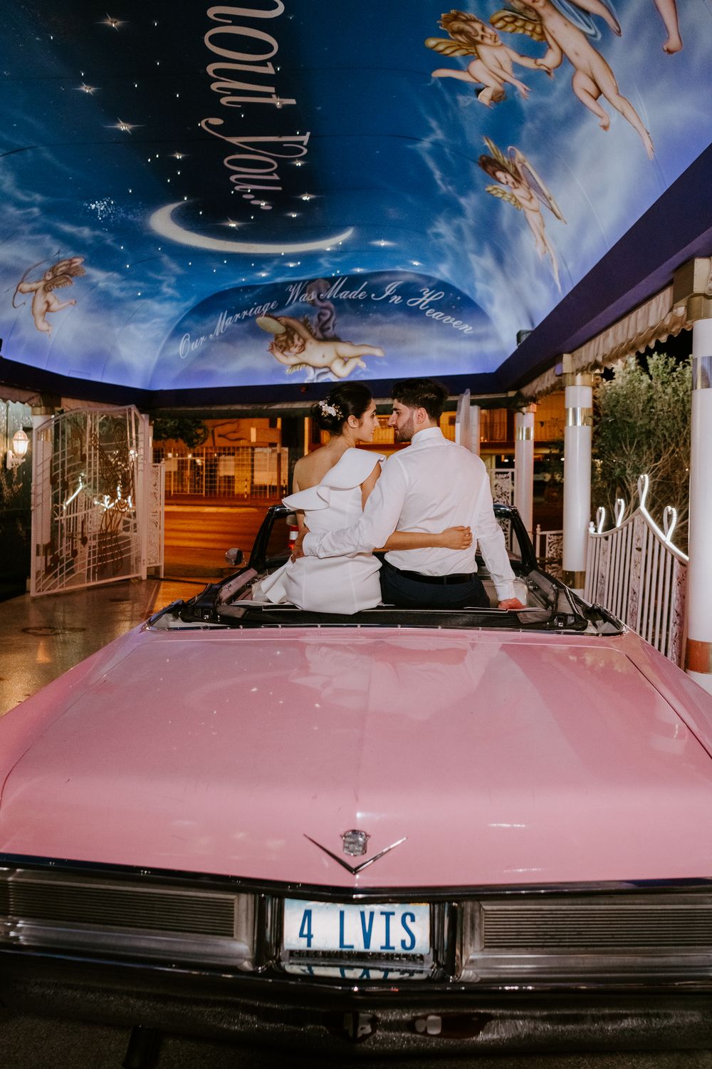 Newlyweds kissing in pink convertible Cadillac under celestial mural at Vegas chapel.