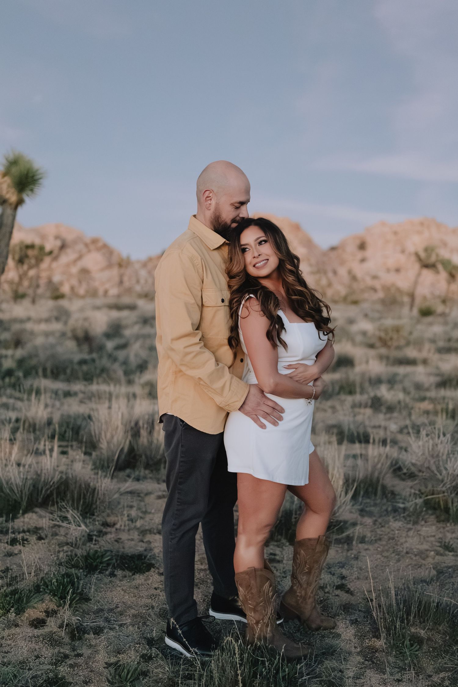A couple poses romantically in Joshua Tree National Park, surrounded by desert shrubs and boulders.
