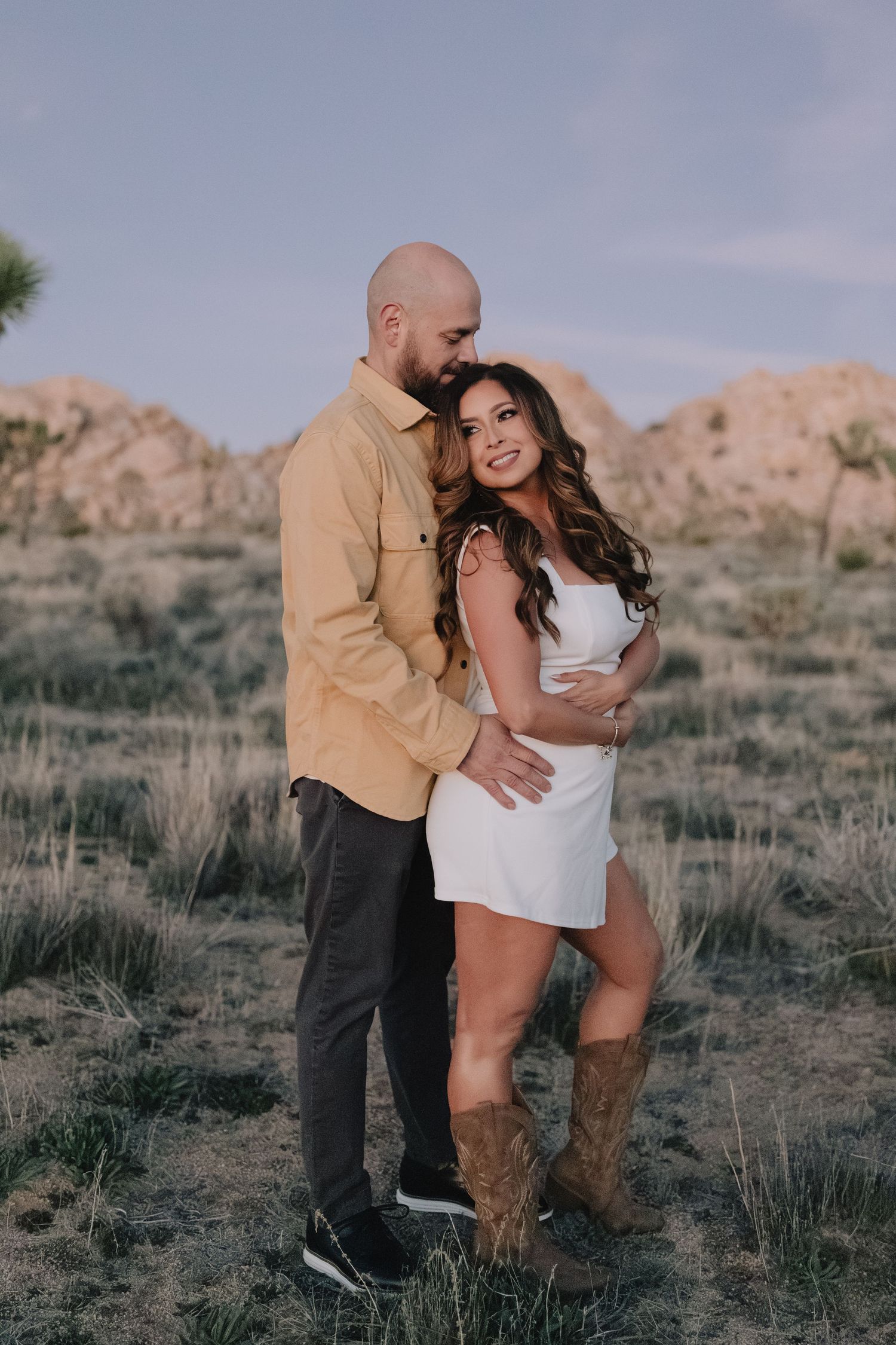 A couple poses romantically in Joshua Tree National Park, surrounded by desert shrubs and boulders.
