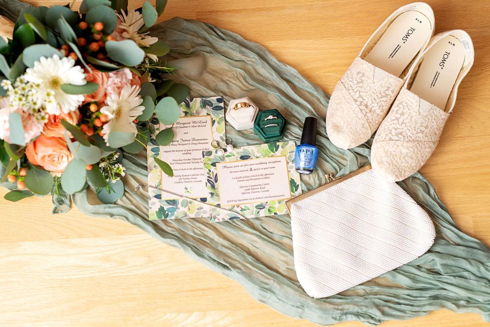 Flat lay of wedding accessories including blush shoes, flowers, and teal fabric on wooden surface.