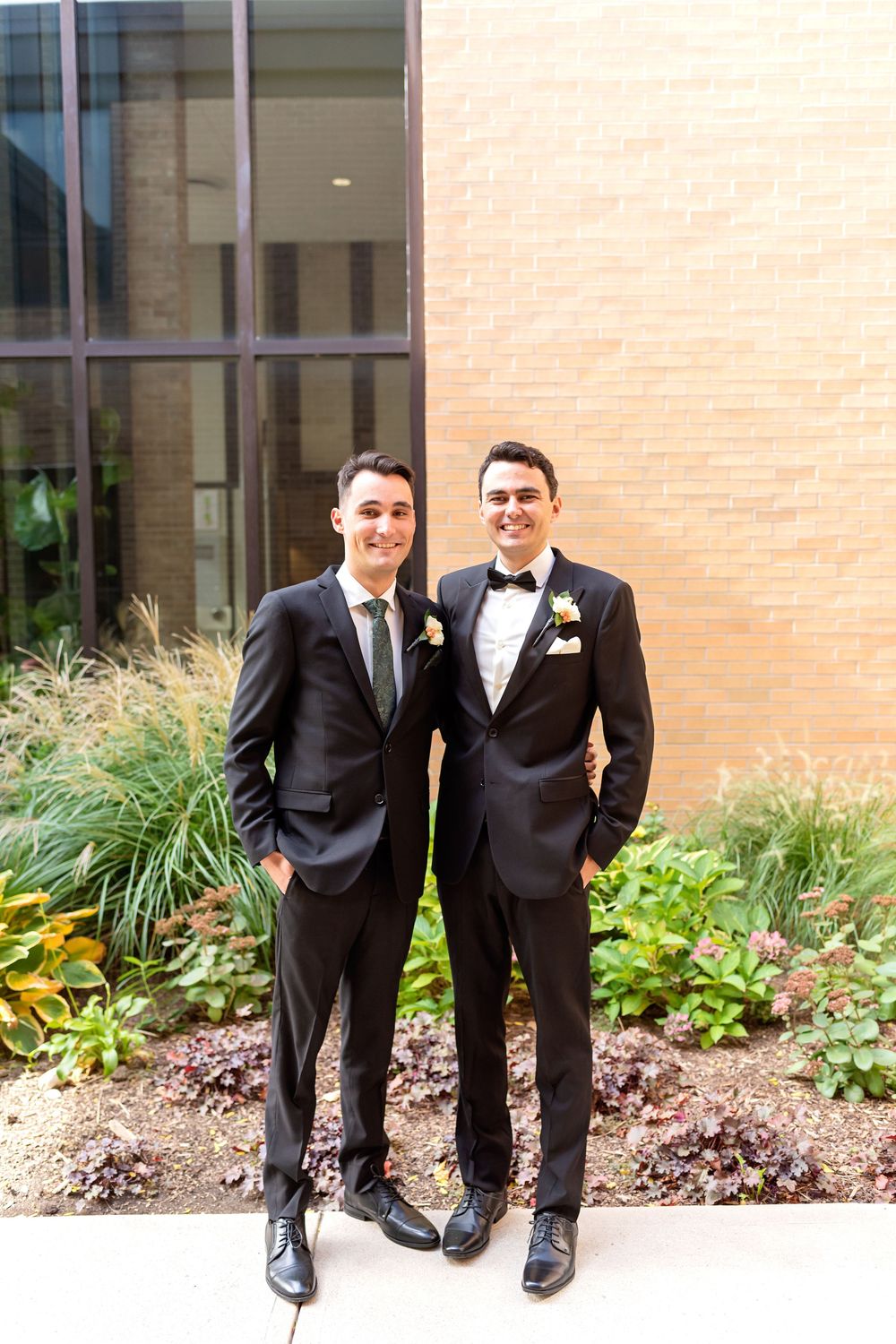 Two people in black tuxedos stand together smiling in front of a brick building with landscaping.