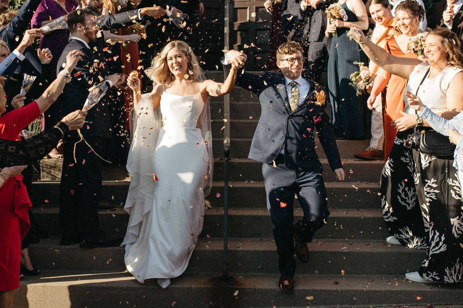 A bride and groom exit their wedding ceremony down steps while guests throw confetti in celebration.
