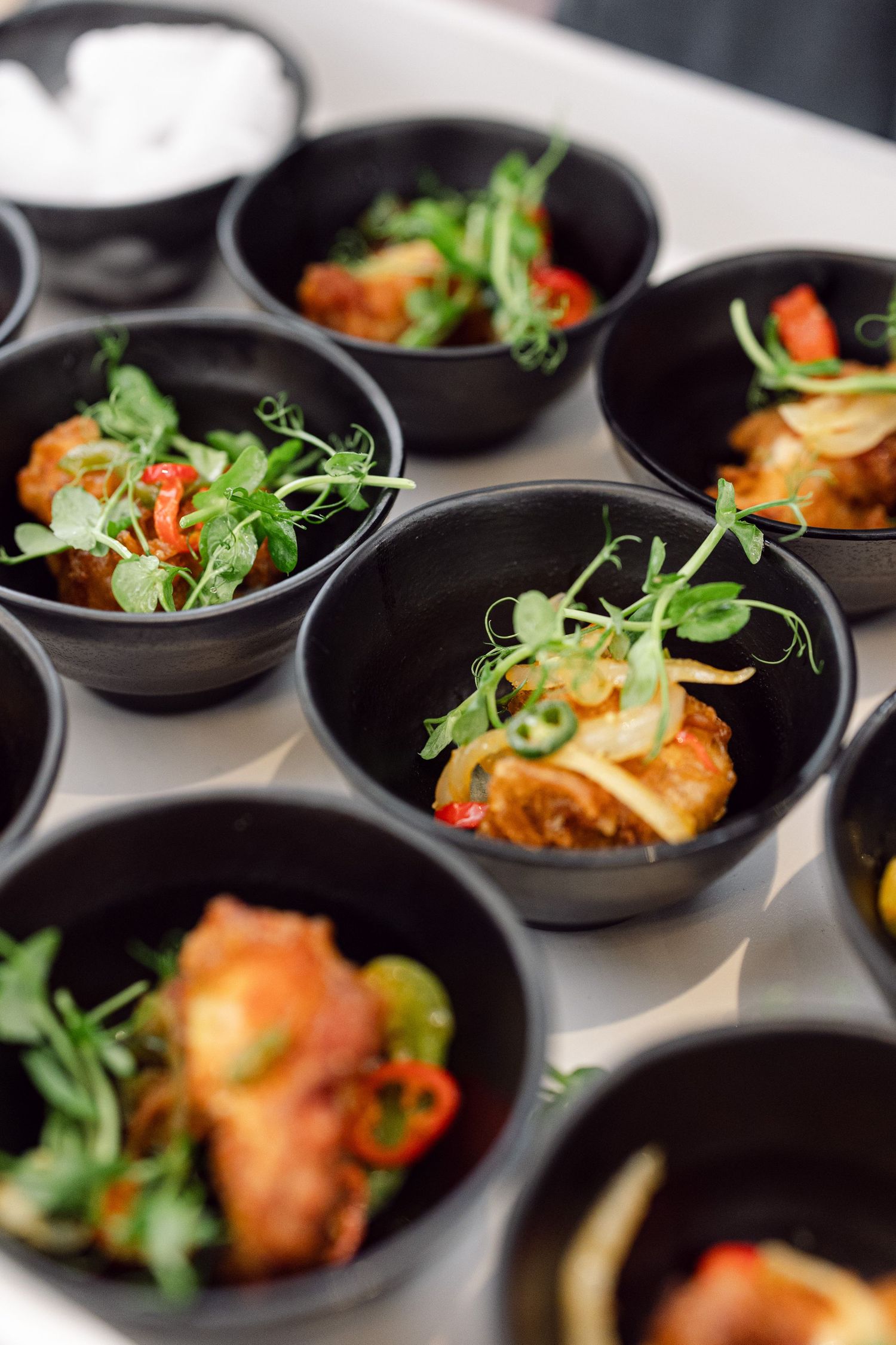 Small black serving bowls filled with crispy shrimp tempura garnished with fresh microgreens on a white surface.