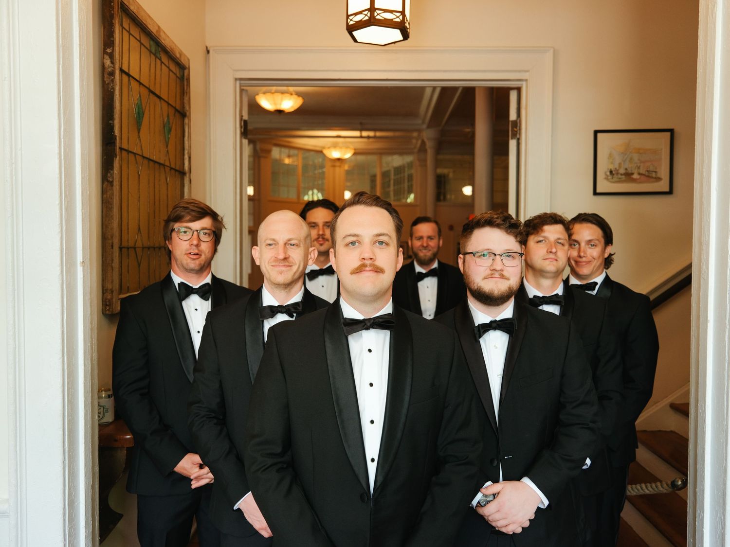 A large group of formally dressed people in tuxedos and black tie attire stand together in an elegant hallway.