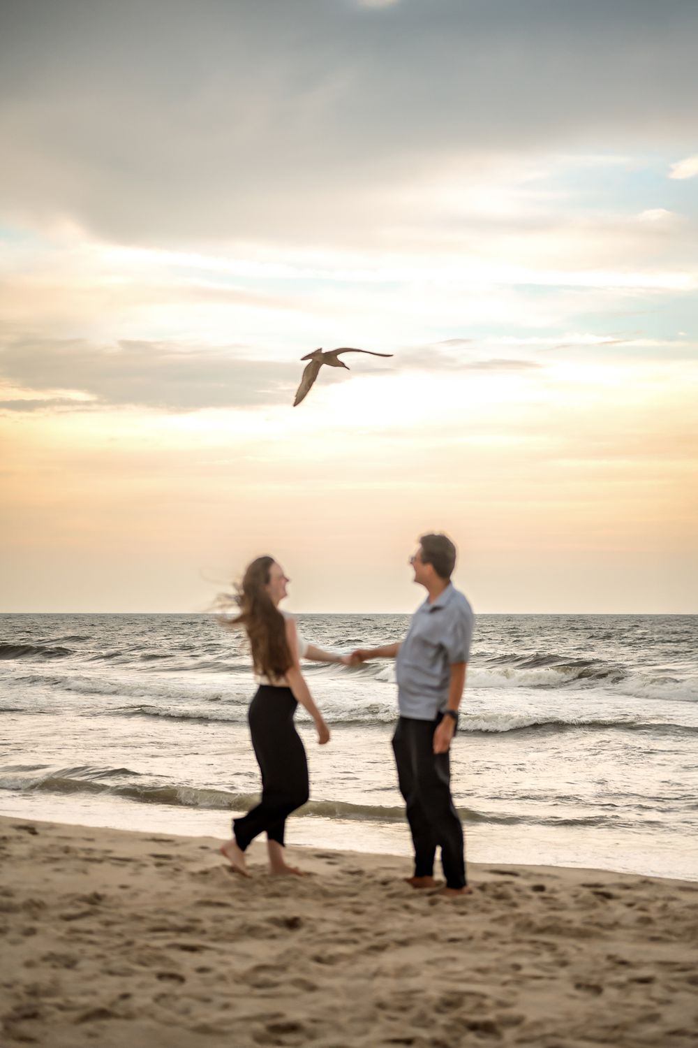 A couple shares a romantic moment on a sandy beach at sunset as seabirds fly overhead.