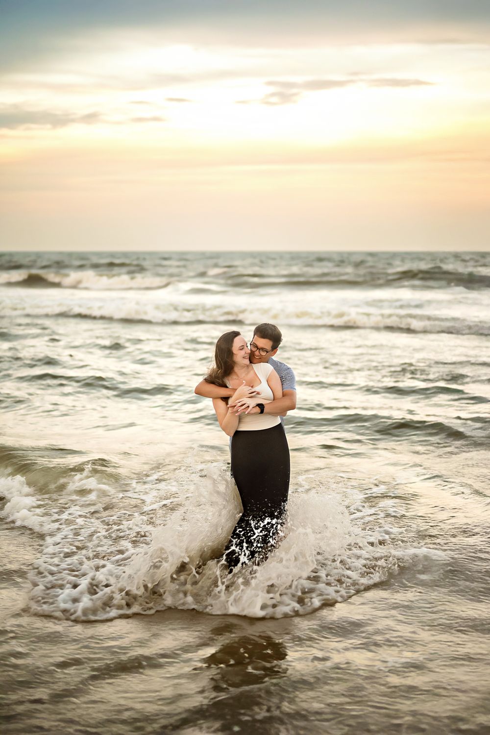 A couple shares an intimate moment while standing in the ocean waves during a golden sunset.