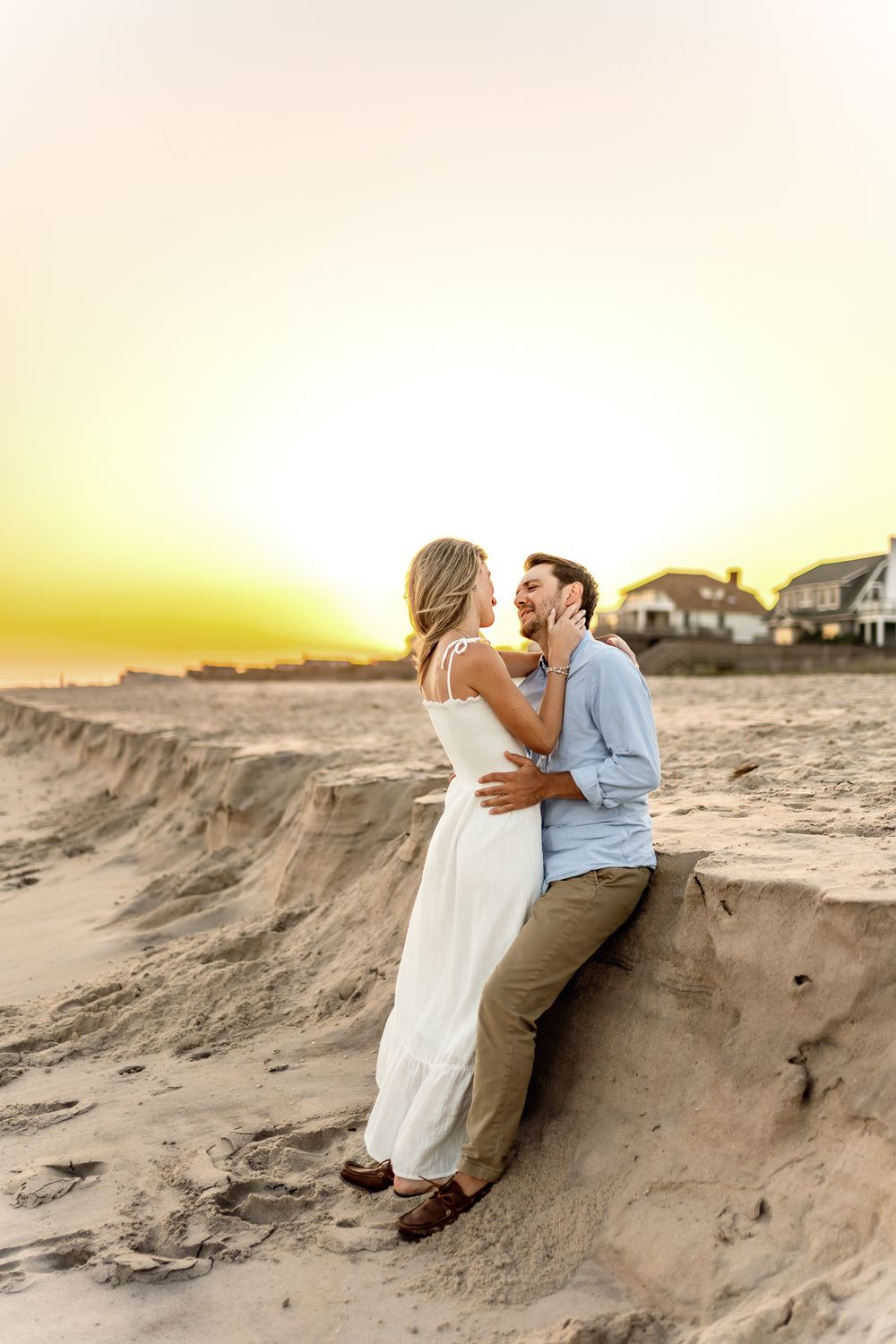 A romantic couple embraces on a sandy beach dune at sunset with beach houses visible in the background.