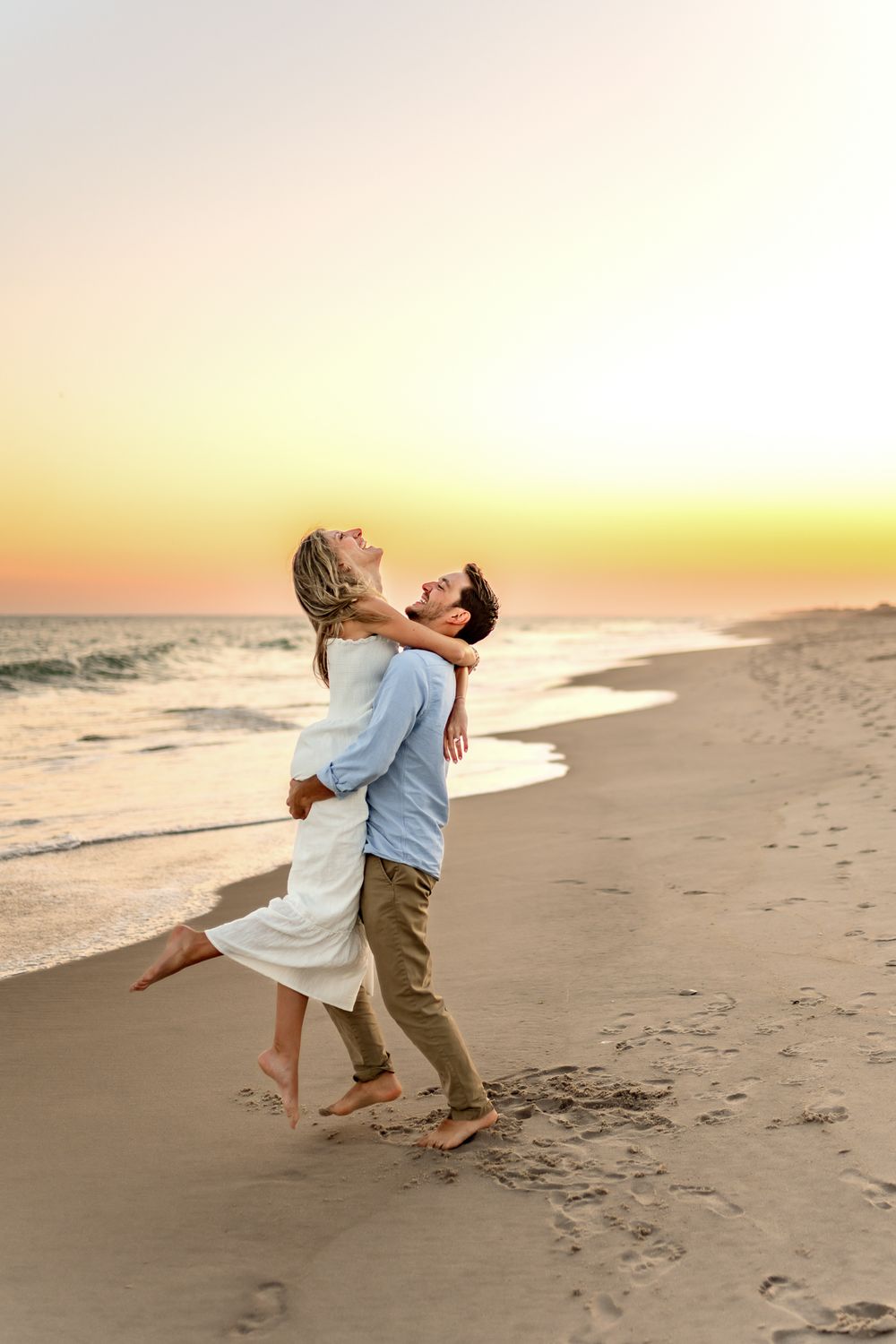 A romantic couple embraces on a sandy beach at sunset with golden light illuminating their joyful moment.