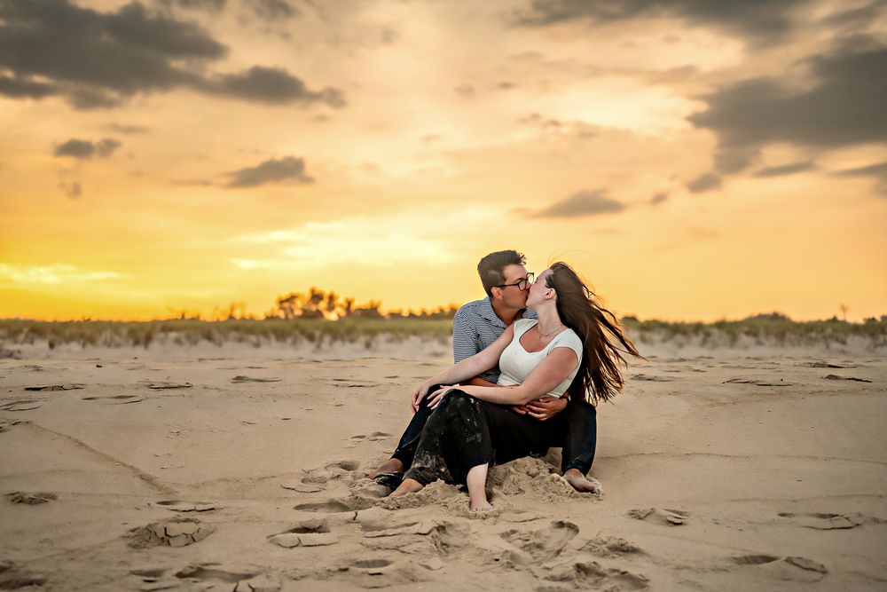 Couple embraces on sandy beach during golden sunset with dramatic cloudy sky in background.