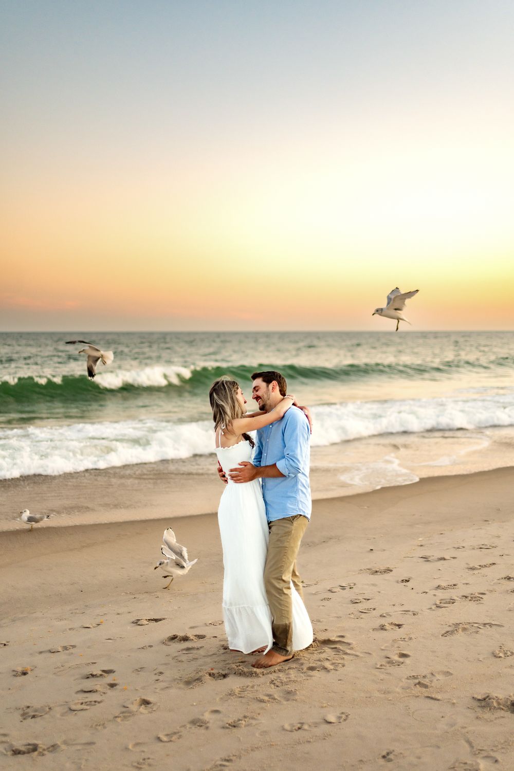 Couple sharing a romantic moment on a sandy beach at sunset as seagulls fly by in the background.
