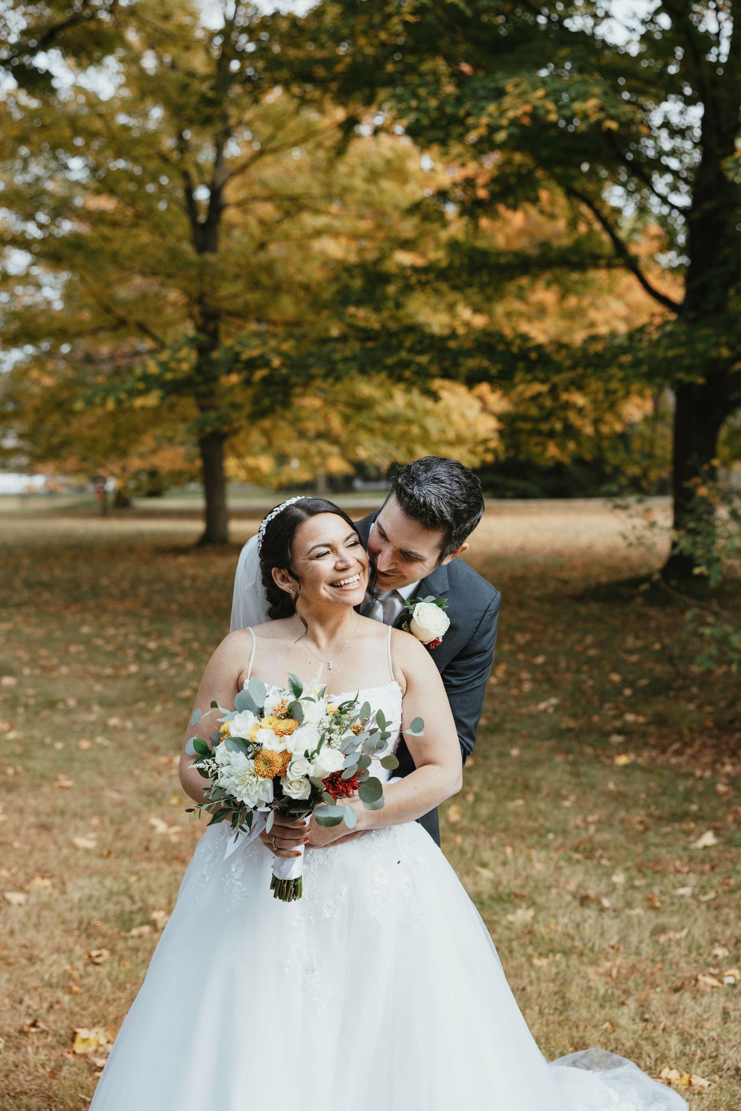A romantic autumn wedding scene with a couple embracing outdoors among fall foliage while holding a floral bouquet.