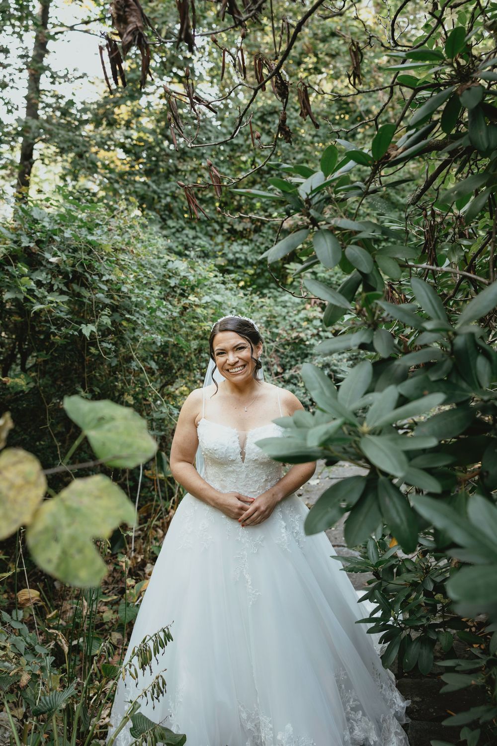 Beautiful bride in a white lace ballgown poses among lush green foliage in an outdoor garden setting.