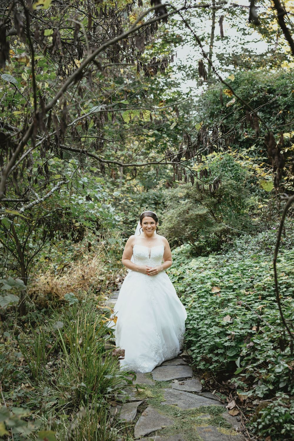 A person in a flowing white wedding dress poses on a stone path surrounded by lush greenery in a garden setting.