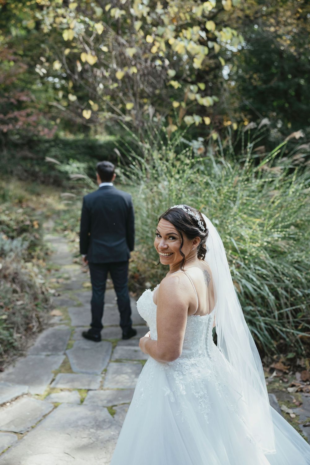 A bride in a white lace wedding dress and veil walks down a garden path while a suited figure stands ahead.
