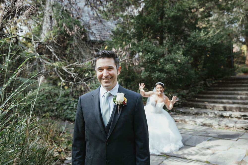 A wedding couple poses on stone steps in a garden setting, with the bride playfully peeking out behind the groom.