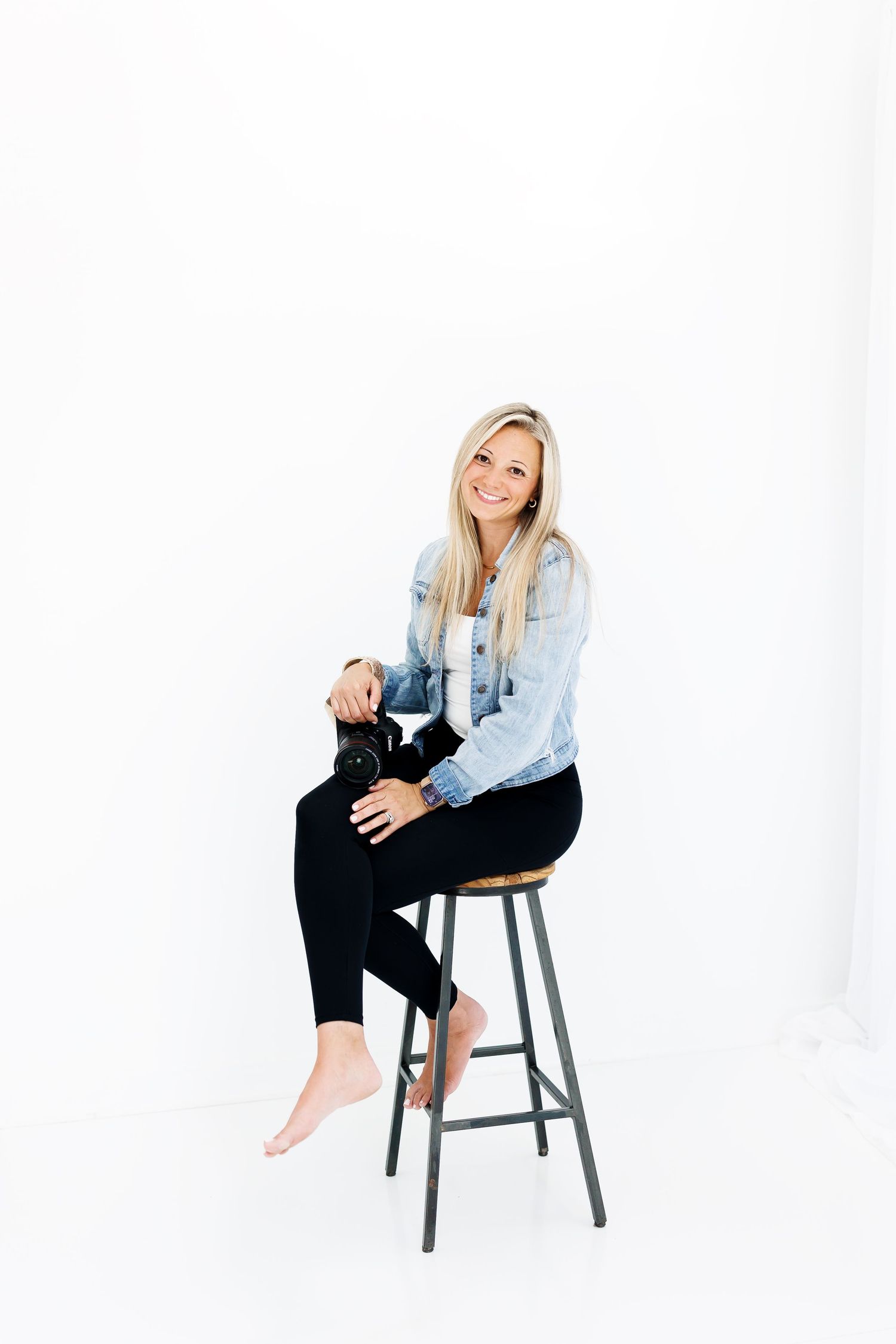 Woman in light denim jacket and black pants poses casually on gray stool against white background in studio setting.