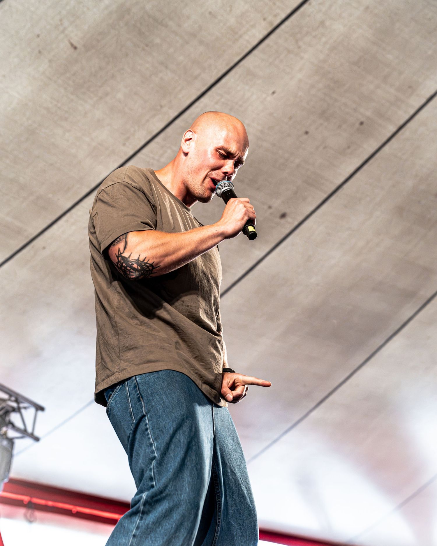 Bald male performer singing into microphone on stage wearing olive t-shirt and jeans with arm tattoos.
