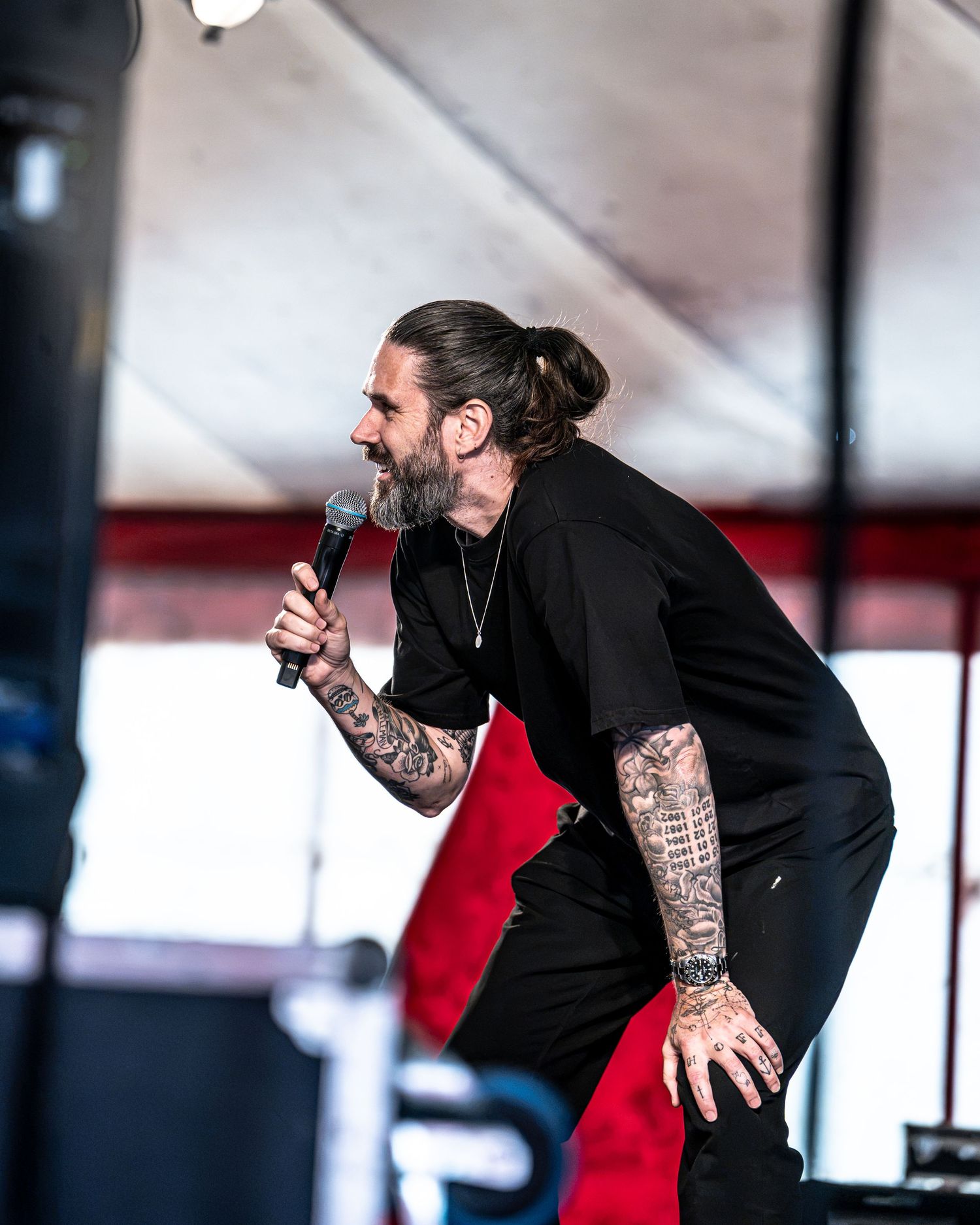 Bearded male vocalist with man bun and full sleeve tattoos performing on stage in black outfit.