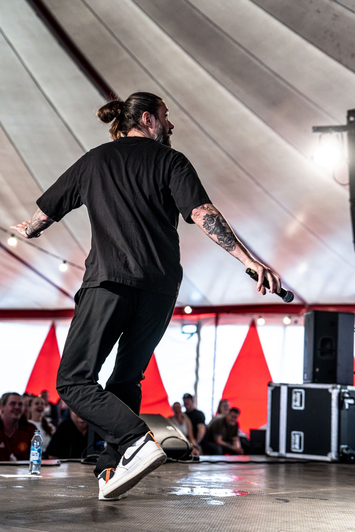 Tattooed performer in black outfit holds microphone on stage at outdoor festival tent venue.