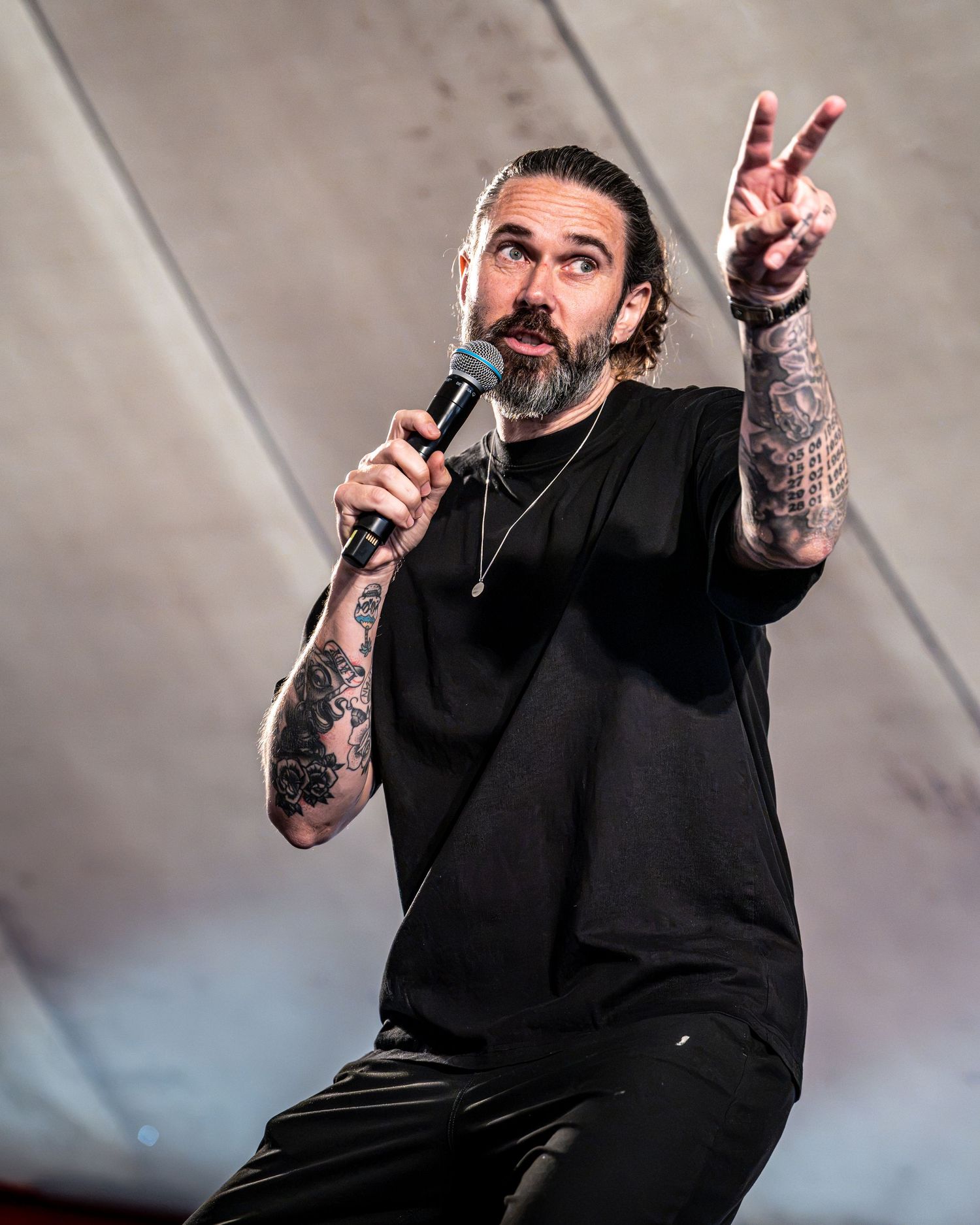 Tattooed man in black t-shirt performing on stage, holding microphone and gesturing with hand raised.