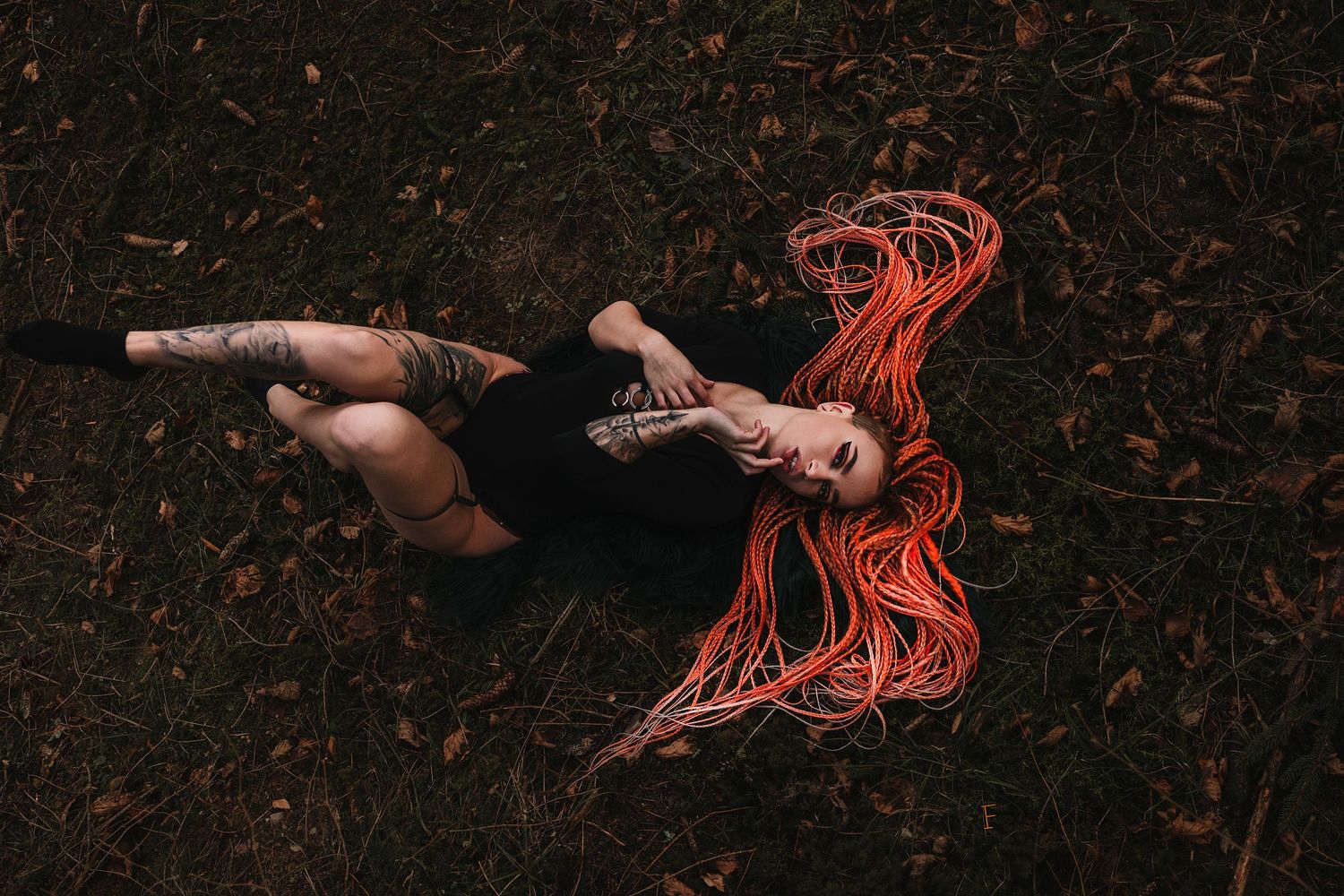 A tattooed woman with long orange braids lies on the ground in a dark forest.