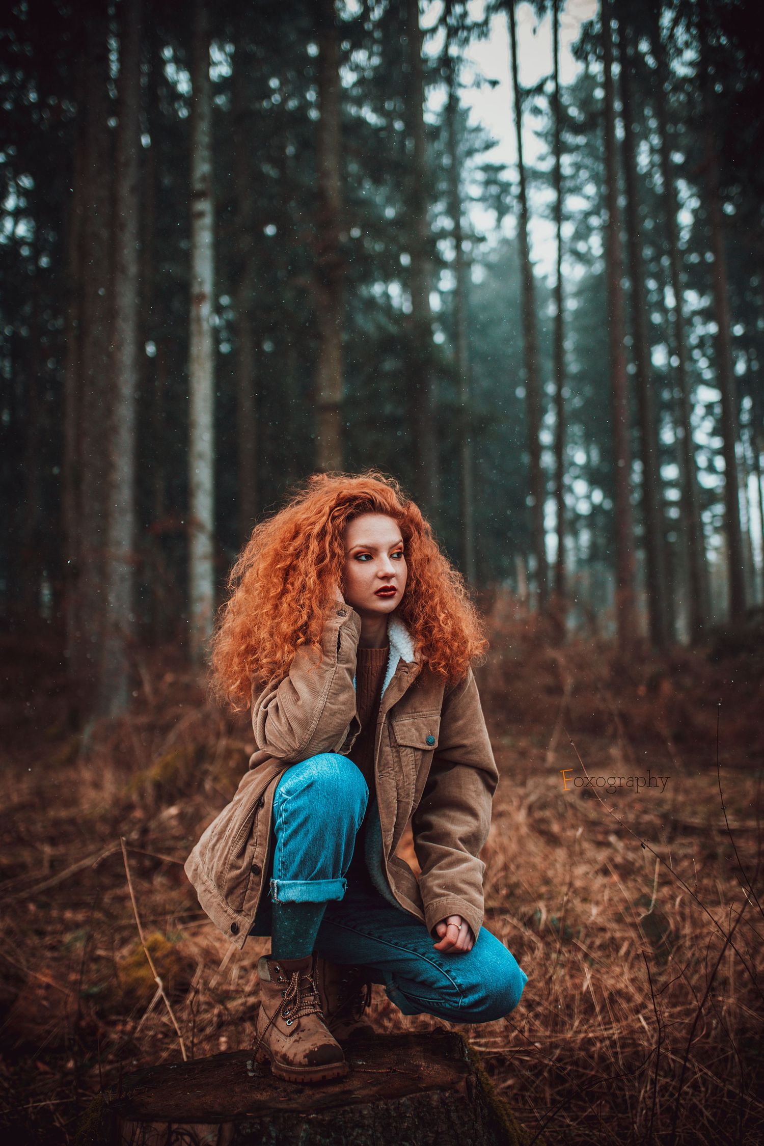 Ryšavá žena v béžovej bunde a modrých džínsoch sedí na pni v borovicovom lese. Umelecká fotografia, Rajecké Teplice.
