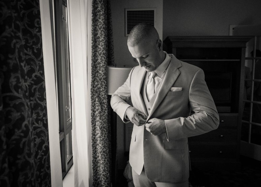 Black and white photo of groom in tan suit buttoning jacket while getting ready by window.