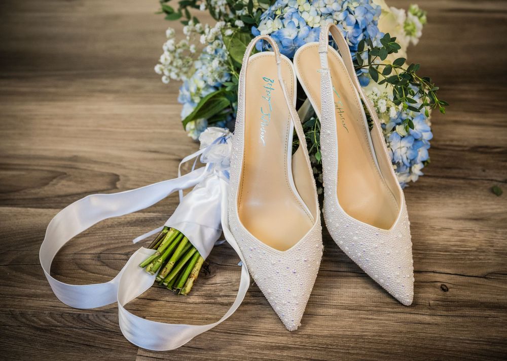 White lace bridal heels with satin ribbon beside blue hydrangea bouquet on rustic wood floor.