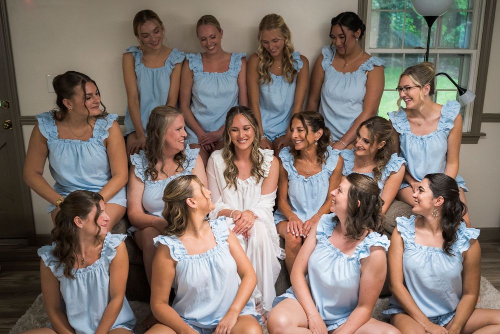 Bride surrounded by bridesmaids in matching light blue tops during wedding preparations indoors.