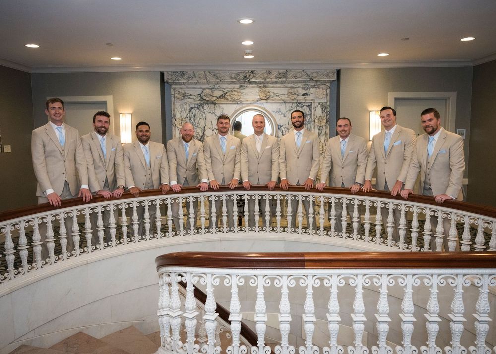 Groomsmen in tan suits posing on a curved staircase balcony at an elegant wedding venue.