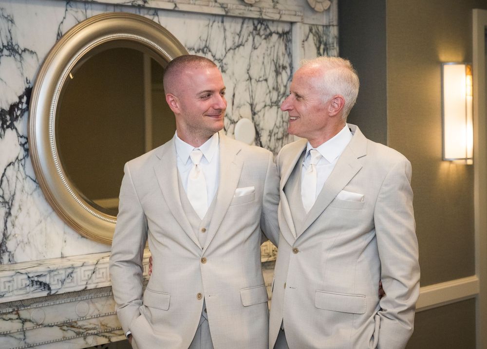 Father and son share a smile in matching tan suits before wedding ceremony near marble fireplace.