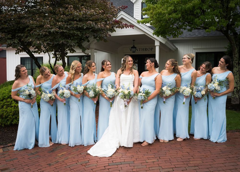 Bride in white gown with bridesmaids in light blue dresses holding white bouquets outdoors.