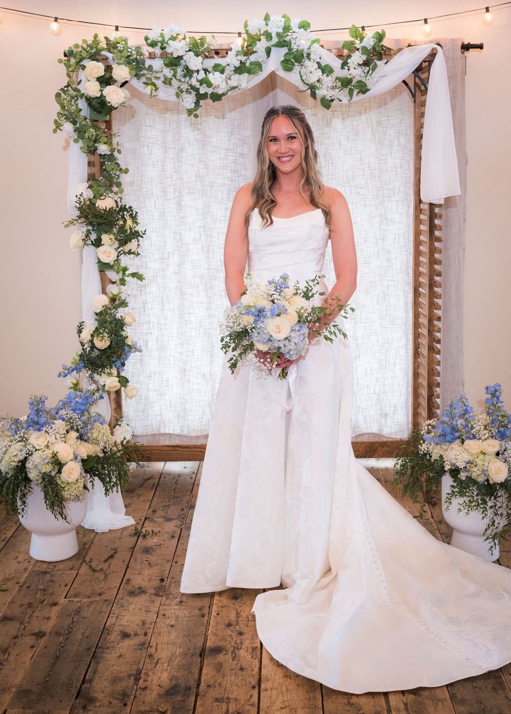 Smiling bride in white strapless gown holding blue and white bouquet standing before floral arch backdrop.